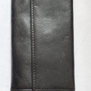 Puede incluir: Un estuche para llaves de cuero negro, plegado y cerrado. De forma rectangular, presenta una costura central. El cuero ligeramente texturizado tiene un acabado mate. Diseñado para guardar llaves, es un accesorio práctico.