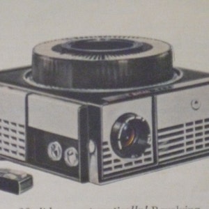May include: Vintage Kodak slide projector with a black and silver design. The projector has a circular top, a lens, and a remote control. The Kodak logo is visible on the side. A classic piece of photographic equipment.