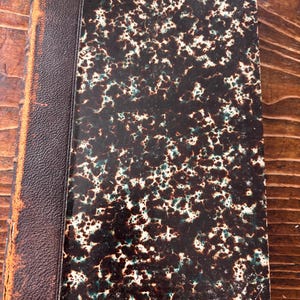 May include: An antique book with a brown leather spine and a patterned cover. The cover features a dark brown and white speckled design with hints of teal. The edges of the book show signs of wear, suggesting its age.