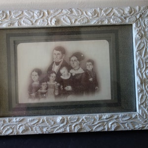 Może przedstawiać: Antyczne czarno-białe zdjęcie rodziny w białej, ozdobnej ramie. Zdjęcie przedstawia mężczyznę, kobietę i pięcioro dzieci.