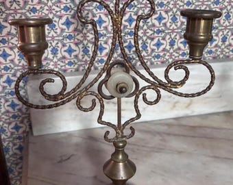 Puede incluir: Un candelabro de latón vintage con tres candeleros. El candelabro tiene un diseño ornamentado con un tallo central y un acento blanco de forma ovalada. La base es redonda y la altura total es de aproximadamente 30 cm.