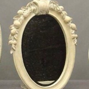 Pode incluir: Um espelho oval branco sujo com um design floral e de folhas decorativo ao redor da moldura. O espelho tem uma superf&iacute;cie escura e reflexiva e um suporte na parte de tr&aacute;s. A moldura tem uma apar&ecirc;ncia texturizada e ornamentada.