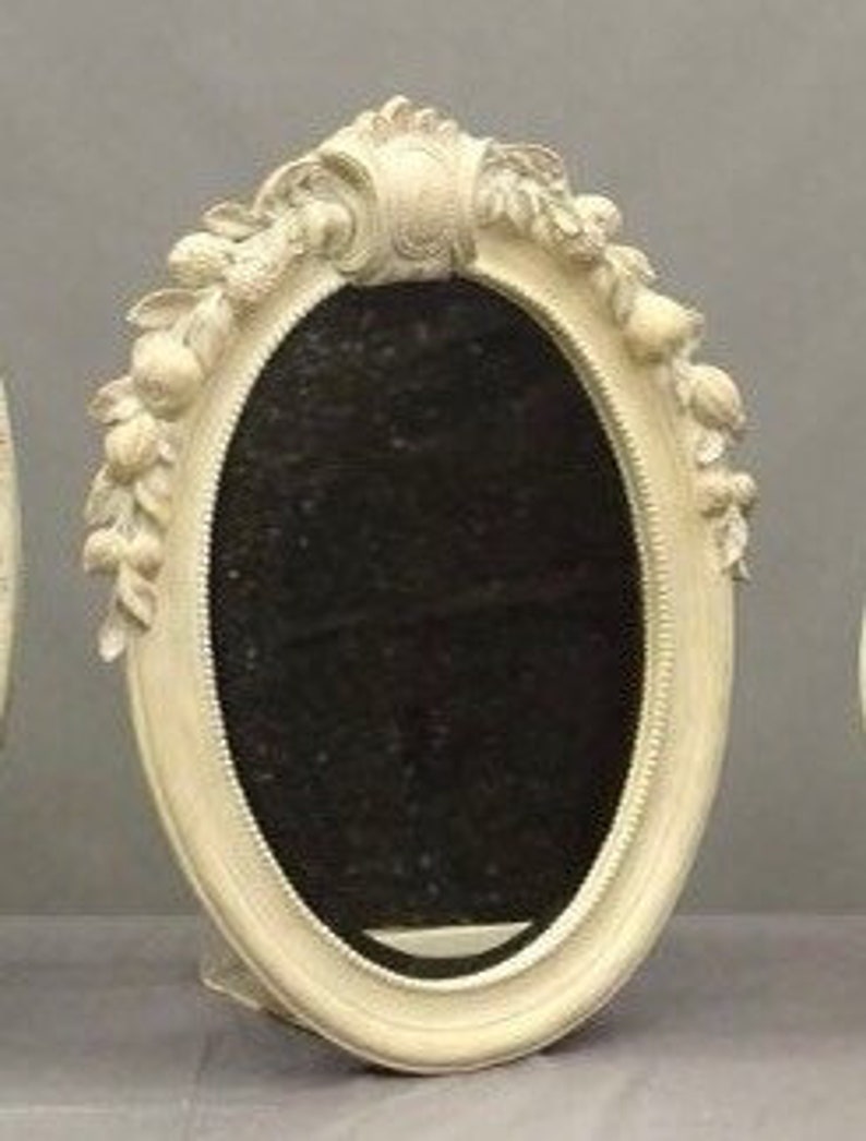 Pode incluir: Um espelho oval branco sujo com um design floral e de folhas decorativo ao redor da moldura. O espelho tem uma superf&iacute;cie escura e reflexiva e um suporte na parte de tr&aacute;s. A moldura tem uma apar&ecirc;ncia texturizada e ornamentada.