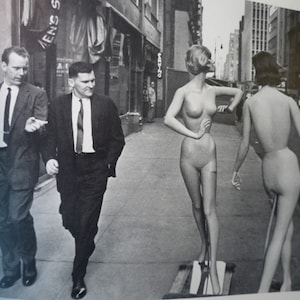 May include: A black and white photo of four people in a city setting. Two men in suits are walking on the sidewalk. Two mannequins are standing in the background. The mannequins are nude and have no clothing on.