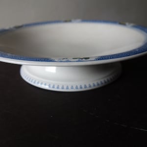 May include: White ceramic pedestal cake stand with a blue floral border. The stand has a wide, shallow bowl and a short, rounded base. The blue border features a repeating floral pattern. A classic piece for displaying desserts.