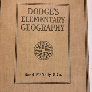 May include: Tan-colored book cover with the title "DODGE'S ELEMENTARY GEOGRAPHY" in black text. A compass rose graphic is centered below the title. The publisher's name, "Rand McNally & Co.", is printed at the bottom.
