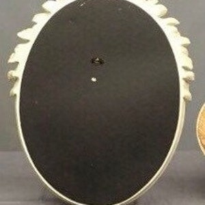 Pode incluir: Parte traseira de uma moldura ovalada creme com uma borda floral decorativa. A moldura tem um fundo preto e um pequeno orif&iacute;cio para pendurar. A moldura &eacute; feita de um material resistente.