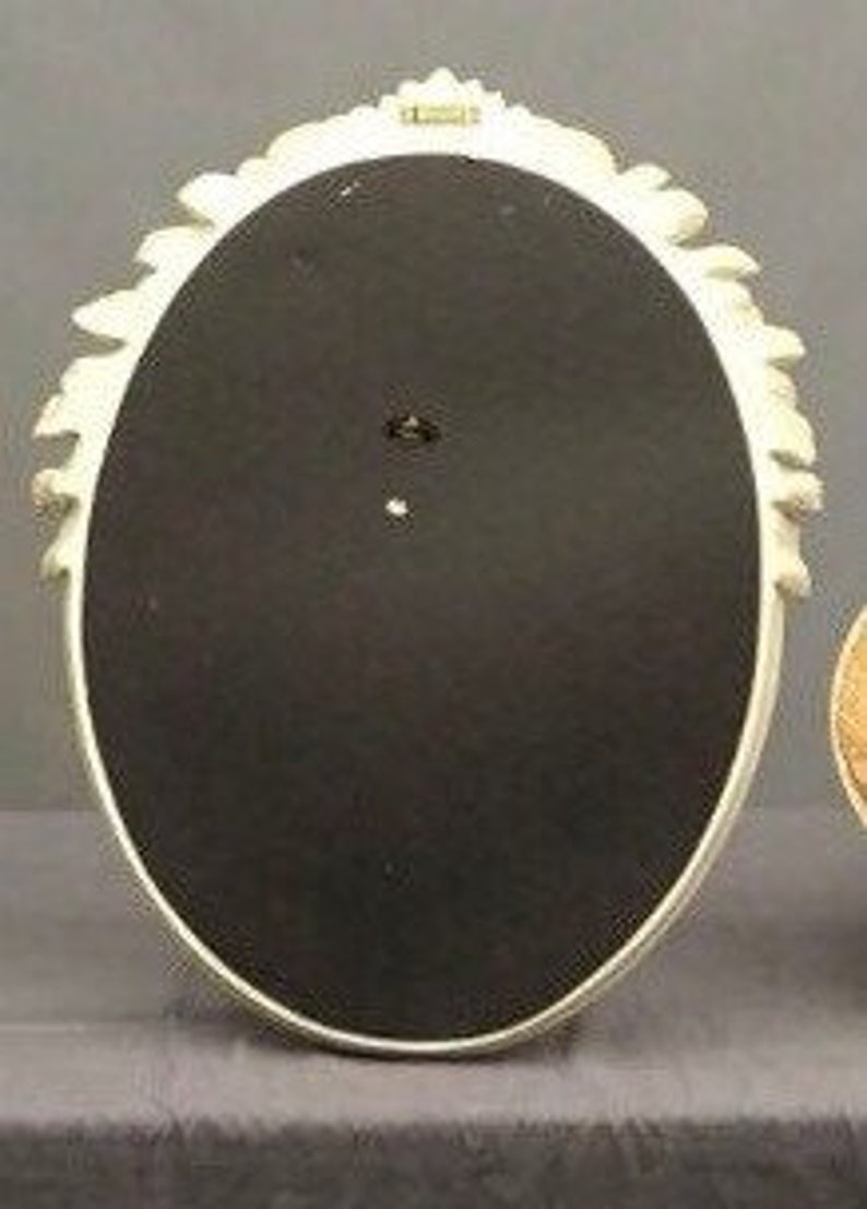Pode incluir: Parte traseira de uma moldura ovalada creme com uma borda floral decorativa. A moldura tem um fundo preto e um pequeno orif&iacute;cio para pendurar. A moldura &eacute; feita de um material resistente.