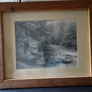 May include: A black and white photograph of a rocky stream in a wooded area, framed in a rustic wooden frame.