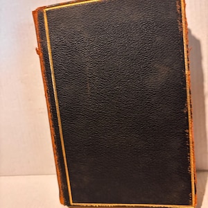 Może przedstawiać: Stara książka z czarną skórzaną okładką i złotymi obramowaniami. Krawędzie książki są w kolorze wyblakłego pomarańczu. Książka jest lekko pochylona, ukazując jej wiekowy wygląd i klasyczny design.