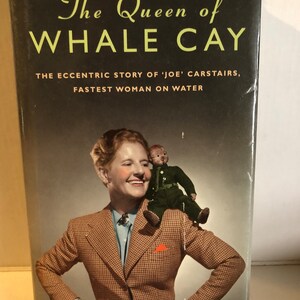 May include: A book cover featuring a photograph of a woman in a checked jacket with a doll on her shoulder. The title of the book is "The Queen of Whale Cay" and the subtitle is "The Eccentric Story of 'Joe' Carstairs, Fastest Woman on Water". The author is Kate Summerscale.