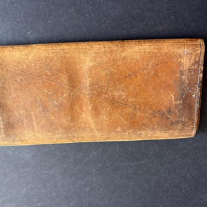May include: A rectangular, well-worn leather wallet with a reddish-brown hue. The wallet shows signs of age with visible scratches and wear, and a red edge. The wallet is closed and appears to be empty.