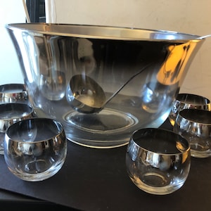 May include: A silver-rimmed glass punch bowl with a matching ladle and eight smaller glasses. The bowl is clear glass with a silver-colored reflective surface. The glasses are also clear glass with a silver rim.