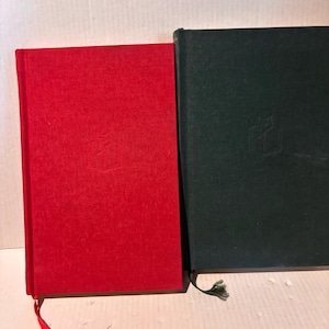 以下が含まれることがあります： 赤と濃い緑の2冊のハードカバーのジャーナルが並んで表示されています。各ジャーナルは、表紙にエンボス加工のデザインと、お揃いのリボンブックマークが特徴です。赤いジャーナルには赤いタッセル、緑のジャーナルには緑のタッセルが付いています。