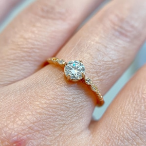 May include: A gold ring featuring a central round brilliant-cut white gemstone held in a four-prong setting. The band is crafted from yellow gold and is accented with small, round, and marquise-shaped white stones along the shank.