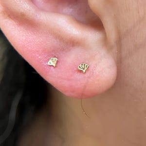 May include: Two small gold stud earrings shaped like ginkgo leaves are positioned on an earlobe. Each earring features a polished metallic finish with delicate, etched lines radiating from the base to the fan-shaped edge of the leaf design.