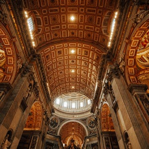 Op de afbeelding: Een uitzicht op het interieur van een grote kathedraal met een hoog gewelfd plafond en sierlijke decoraties. Het plafond is gemaakt van hout en heeft een patroon van vierkanten en rechthoeken. De muren zijn van steen en hebben kolommen en bogen. In het midden van de ruimte staat een groot altaar.