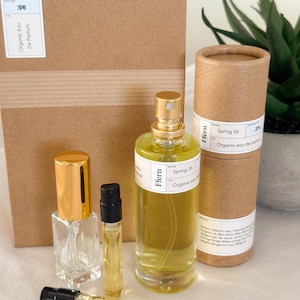 May include: A collection of perfume bottles and samples. The main bottle is clear with a gold cap, labeled "Ffern Spring 26 Organic eau de parfum." Additional samples and a cylindrical container with the same label are also present. A cardboard box is in the background.