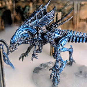 May include: A detailed, metallic blue figurine of an Alien Queen. The creature has a long, segmented tail, multiple limbs, and a menacing head with sharp teeth. The figure is posed in a dynamic stance, with intricate detailing.