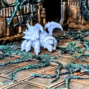 May include: A light gray, unpainted miniature creature figurine featuring a segmented, armored exoskeleton, multiple legs, and sharp, curved appendages, positioned on a textured, metallic-looking floor with dark green, vine-like growths.