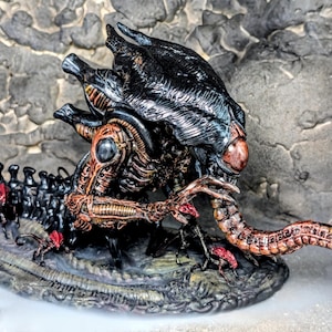 May include: A detailed sculpture of an Alien creature, featuring a black, textured head and a segmented body. The figure has a long, segmented tail and is posed on a textured base. The color palette includes black, bronze, and red accents.