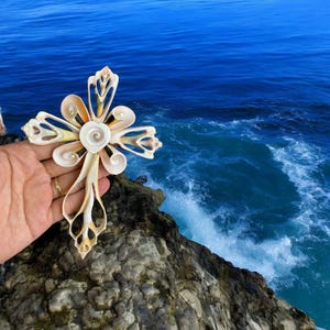 May include: A handcrafted cross made from seashells, featuring a central spiral design and intricate petal-like shapes. The cross is held against a backdrop of a deep blue ocean and rocky cliffs, showcasing natural textures and colors.