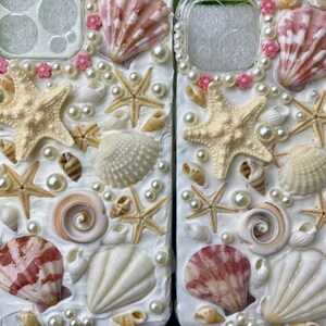 May include: Two white phone cases decorated with seashells, pearls, and starfish. The cases have a white background and are covered in a variety of seashells, pearls, and starfish. The shells are in shades of white, pink, and brown. The pearls are white and the starfish are white.
