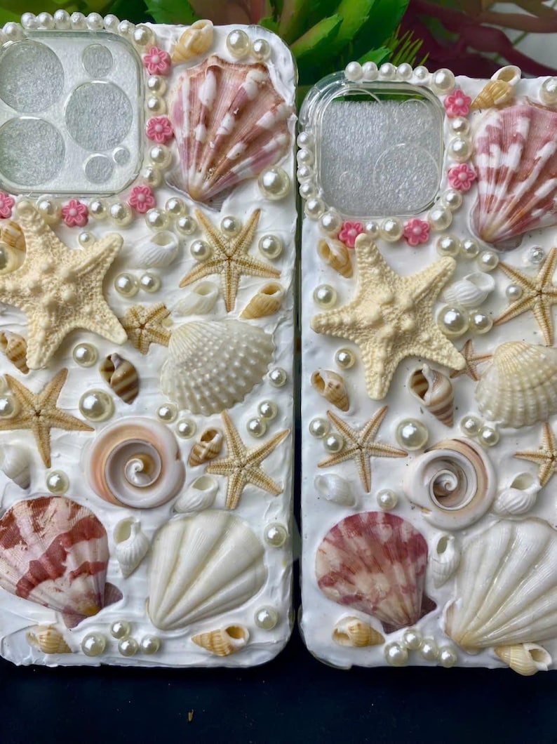 May include: Two white phone cases decorated with seashells, pearls, and starfish. The cases have a white background and are covered in a variety of seashells, pearls, and starfish. The shells are in shades of white, pink, and brown. The pearls are white and the starfish are white.