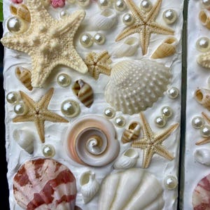 May include: A white phone case decorated with seashells, starfish, and pearls. The case has a textured white background and features a variety of seashells in different sizes and colors.