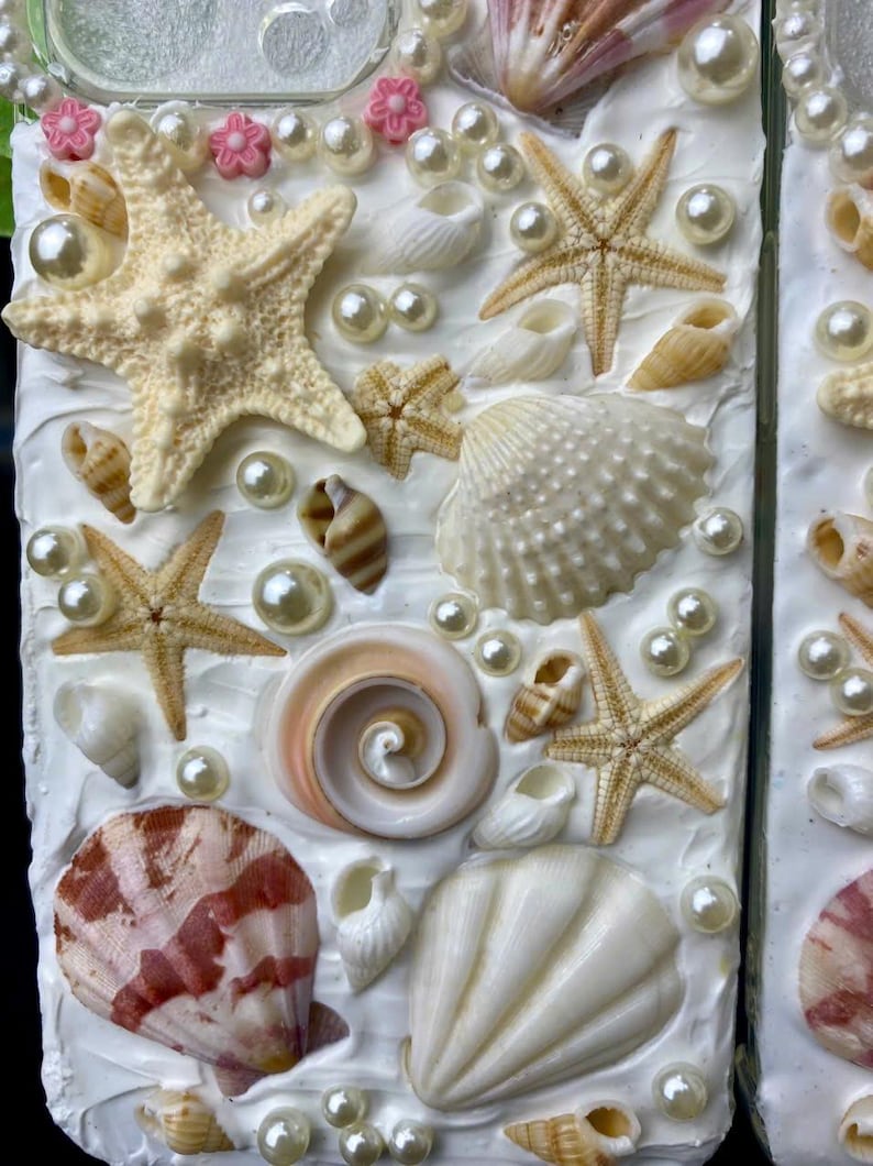 May include: A white phone case decorated with seashells, starfish, and pearls. The case has a textured white background and features a variety of seashells in different sizes and colors.