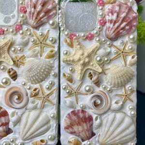 May include: Two white phone cases decorated with seashells, pearls, and starfish. The cases have a clear plastic window for the camera lens.