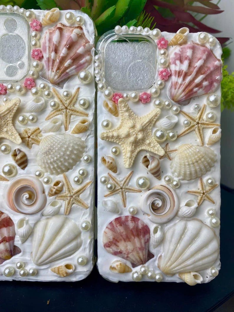 May include: Two white phone cases decorated with seashells, pearls, and starfish. The cases have a clear plastic window for the camera lens.