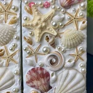 May include: A white phone case decorated with seashells, starfish, and pearls. The case has a pink and white floral design around the camera lens.