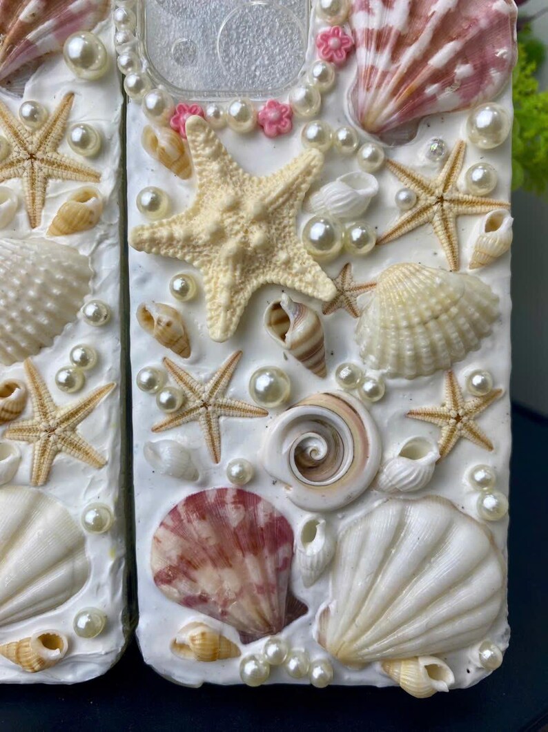 May include: A white phone case decorated with seashells, starfish, and pearls. The case has a pink and white floral design around the camera lens.
