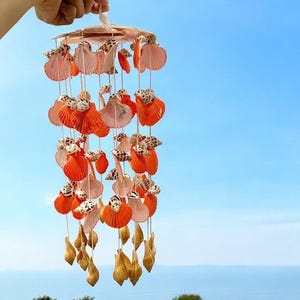 May include: A seashell mobile, perfect for a coastal theme. It features orange and pink seashells, small patterned shells, and dangling golden shells. The mobile hangs against a clear blue sky, evoking a sense of the seaside.