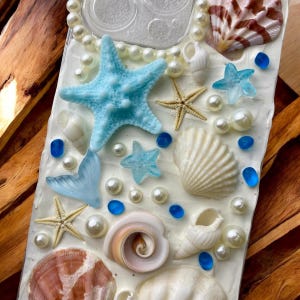 May include: A white phone case decorated with seashells, starfish, pearls, and blue glass beads. The case has a clear back panel and a white border.