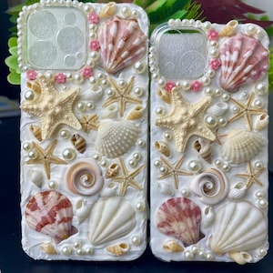 May include: Two white phone cases decorated with seashells, pearls, and pink accents. The cases feature a variety of seashells, including starfish, clams, and spiral shells.