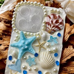 May include: A white phone case decorated with seashells, starfish, pearls, and blue glass beads. The case has a clear back with a cutout for the camera lens.