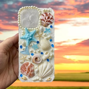 May include: A white phone case decorated with seashells, starfish, and pearls. The case has a blue and white color scheme and features a variety of different seashells.