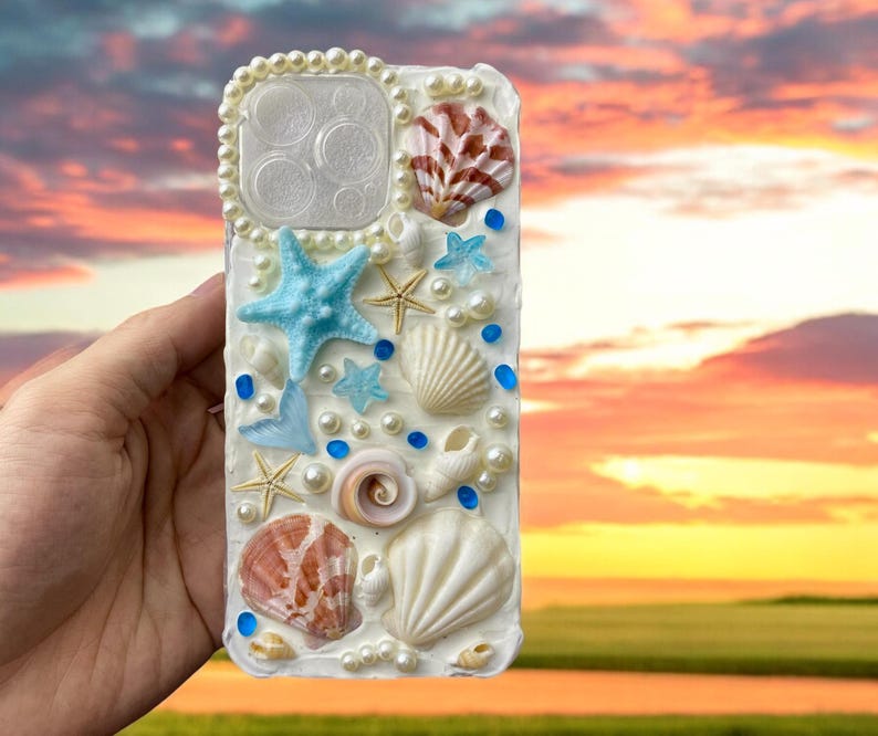 May include: A white phone case decorated with seashells, starfish, and pearls. The case has a blue and white color scheme and features a variety of different seashells.