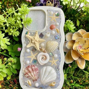 Op de afbeelding: Witte telefoonhoes versierd met schelpen, zeesterren, parels en iriserende accenten. De hoes heeft een verscheidenheid aan schelpen in verschillende vormen en kleuren, waardoor een strandthema ontstaat. Omringd door groen gebladerte.