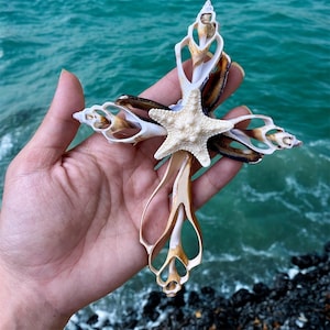 May include: A handcrafted cross made from seashells and a starfish. The cross is held in a hand, with the ocean in the background. The cross is made of various seashells in shades of white, brown, and beige, with a white starfish at the center.
