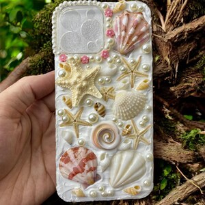 May include: A white phone case decorated with seashells, pearls, and starfish. The case has a clear window for the camera lens.