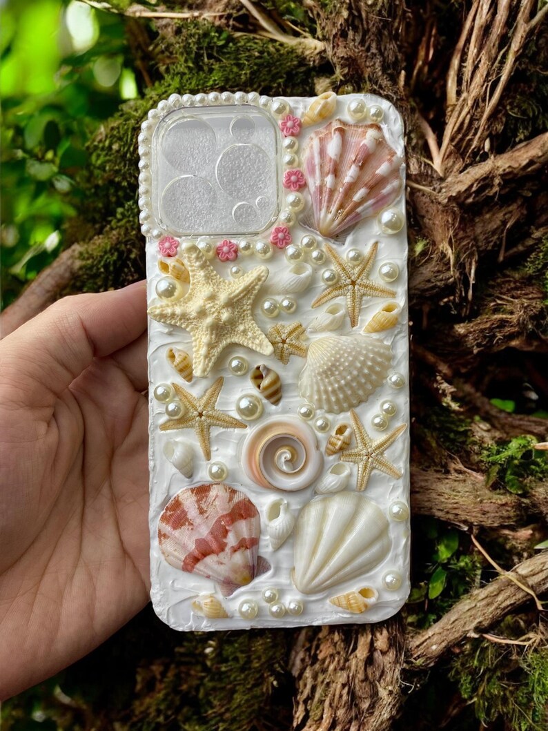 May include: A white phone case decorated with seashells, pearls, and starfish. The case has a clear window for the camera lens.