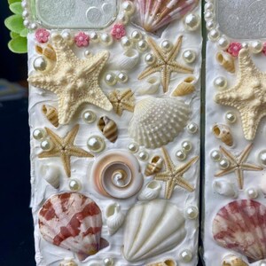 May include: A white phone case decorated with seashells, starfish, and pearls. The case has a clear window on the back.