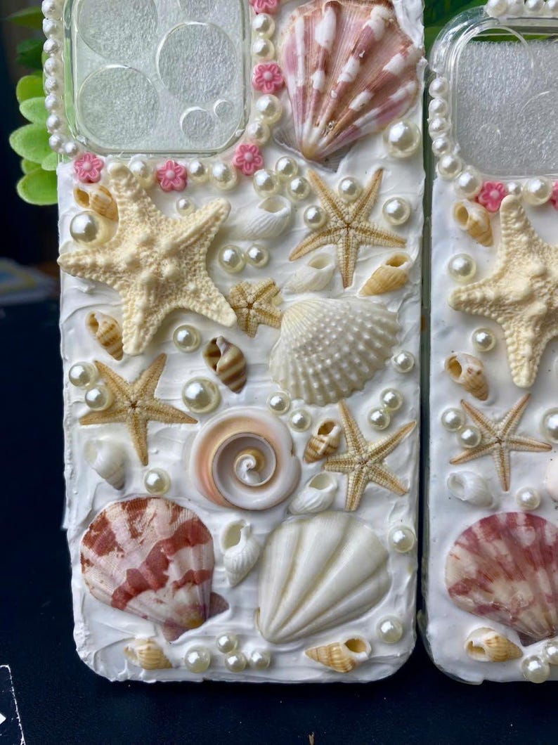 May include: A white phone case decorated with seashells, starfish, and pearls. The case has a clear window on the back.