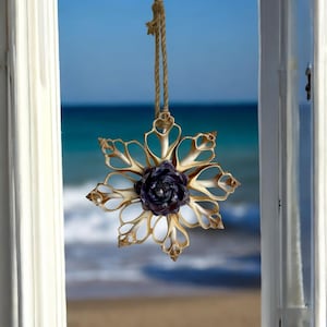 May include: A seashell snowflake ornament with a purple seashell flower in the centre, hanging in a window with a view of the sea.