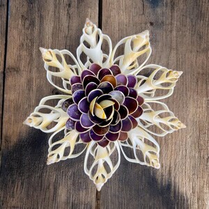 May include: A white seashell flower with a purple and yellow centre. The flower is made of many small seashells arranged in a circular pattern.