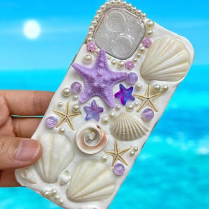 May include: A white phone case decorated with seashells, starfish, pearls, and iridescent beads. The case has a purple starfish in the center and a clear camera lens cover.