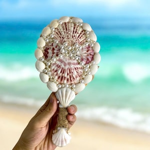 Op de afbeelding: Een handspiegel gemaakt van schelpen en parels. De spiegel is ovaalvormig en heeft een handvat gemaakt van een schelp. De spiegel is versierd met witte parels en schelpen.